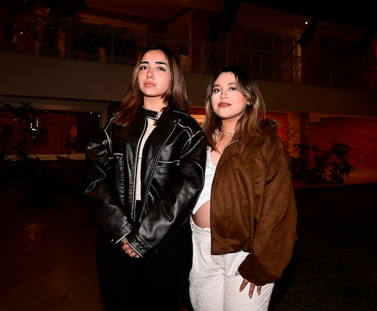 Mujeres jóvenes, una en chaqueta de cuero negra y otra embarazada en chaqueta marrón y blusa blanca, posan sonrientes en evento nocturno.