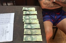 Hombre tatuado, vestido con pantalón corto colorido, sentado con brazos cruzados y billetes sobre la mesa, en ambiente sencillo.