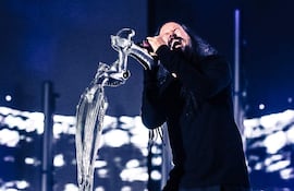 Hombre con cabello largo y oscuro, vestido de negro, canta con intensa concentración en un escenario iluminado.