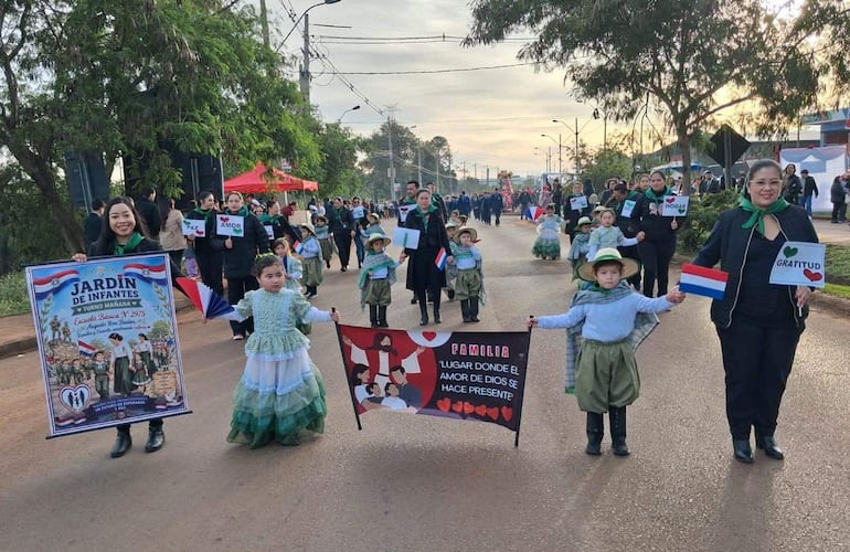 Mujer sostiene cartel de 'Jardín de Infantes' acompañada de niños con trajes típicos, en un ambiente festivo en la calle.