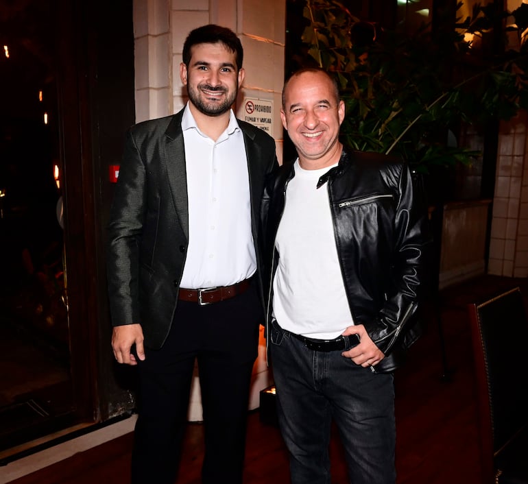 Dos hombres sonríen, uno viste camisa blanca con saco oscuro y el otro camiseta blanca bajo chaqueta de cuero negra en ambiente moderno.