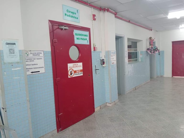 Pasillo del hospital con puerta roja señalando 'Cirugía Pediátrica' y anuncios de distanciamiento social.