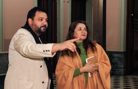 Hombre de traje claro y barba explica algo, mientras una mujer con abrigo beige lo escucha atentamente y toma notas.