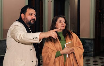 Hombre de traje claro y barba explica algo, mientras una mujer con abrigo beige lo escucha atentamente y toma notas.