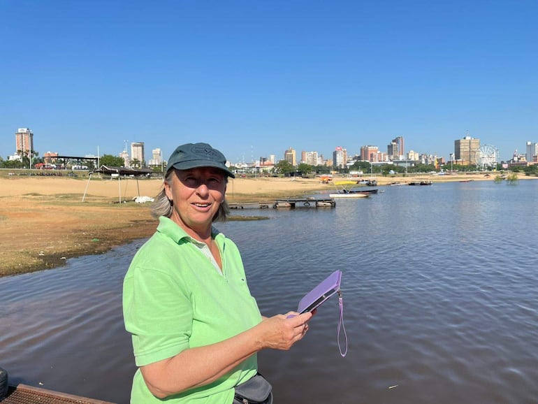 Mujer de cabello largo y canoso, con camiseta verde y gorra gris, sonríe mientras sostiene una tablet junto al río.