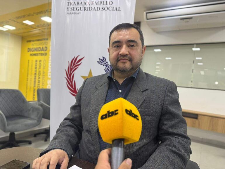 Jorge Rivas con saco gris y camisa de rayas, sostiene un micrófono en sala con logo del MTESS, fondo blanco y sillas grises.