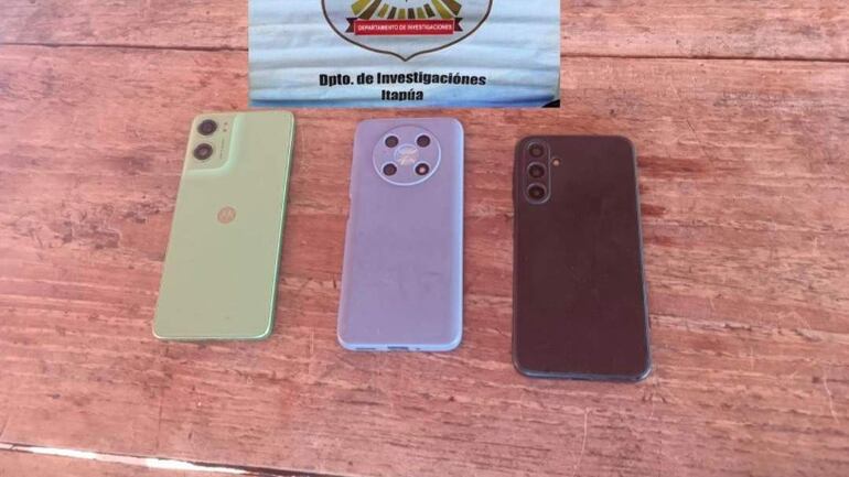 Tres teléfonos móviles en una superficie de madera. Cartel que indica el Dpto. de Investigaciones de Itapúa en el fondo.