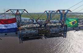 Puente en construcción con banderas de Paraguay y Brasil, en un cielo despejado y sin personas visibles.