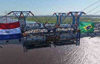 Puente en construcción con banderas de Paraguay y Brasil, en un cielo despejado y sin personas visibles.