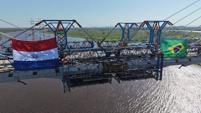 Puente en construcción con banderas de Paraguay y Brasil, en un cielo despejado y sin personas visibles.