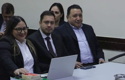 Mujer de cabello lacio y chaqueta oscura sonríe, mientras dos hombres en traje tienen expresiones relajadas, en una mesa de reuniones.