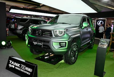Un coches SUV gris metálico con detalles verdes, expuesto en un evento con fondo moderno y personas asistentes difusas.