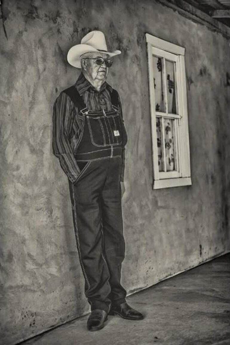 Hombre mayor con sombrero blanco y gafas de sol, apoyado en una pared, luciendo sereno y vestido con overol oscuro y camisa a rayas.