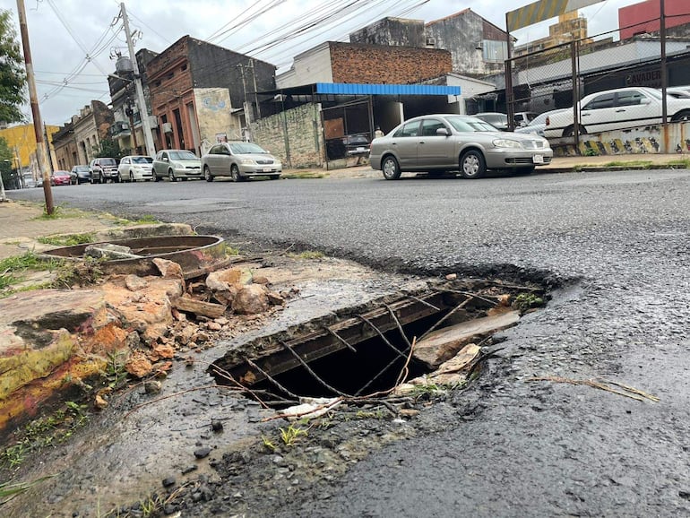 Calle con pavimento dañado, hueco en la vía y vehículos estacionados bajo un cielo nublado.
