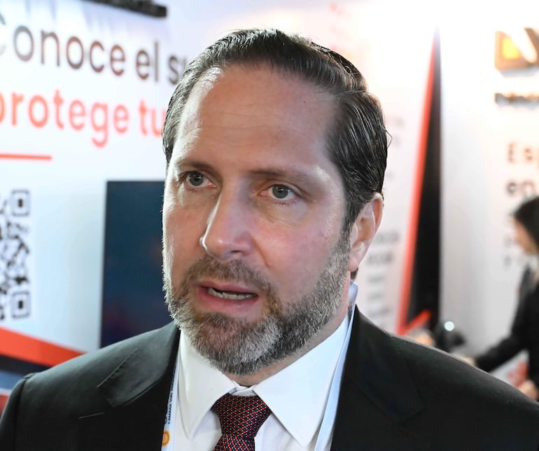 Hombre de mediana edad con barba y corbata roja, hablando en evento con paneles informativos de fondo.