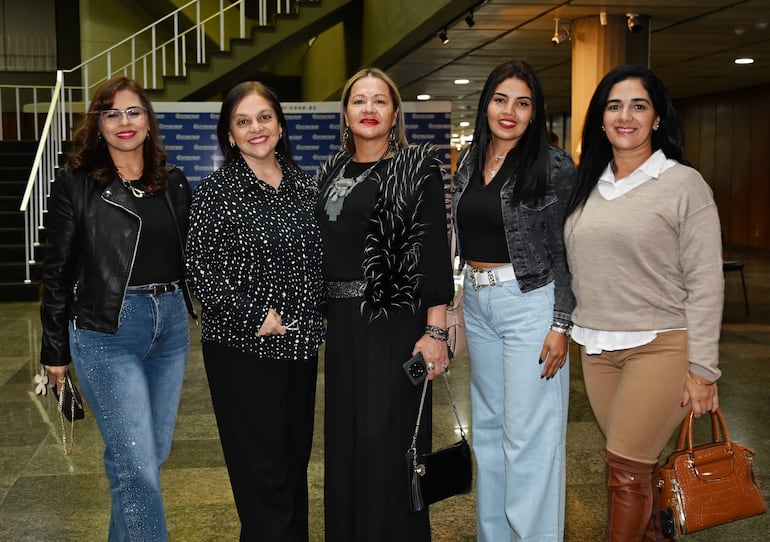 Cinco mujeres posan sonrientes, vestidas elegantemente en tonos oscuros y claros, en un ambiente formal con un banner al fondo.