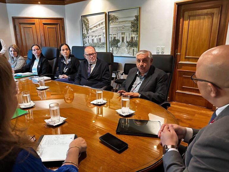 Fretes de cabello canoso habla en reunión, rodeado de persona con atuendo formal, en ambiente de sala de conferencias luminosa.