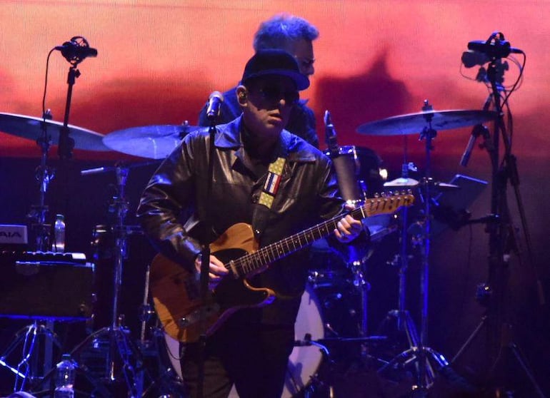 Andrés Calamaro en el escenario con gafas oscuras, sombrero y chaqueta de cuero, tocando guitarra eléctrica con luces de fondo.