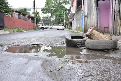 Calle urbana con bache grande y agua, neumáticos desechados y señal de tráfico 'PARE'.