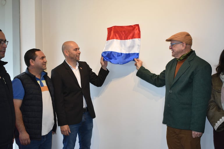 Hombre en traje oscuro sostiene la bandera paraguaya, acompañado de otros hombres, en un ambiente formal y sobrio.