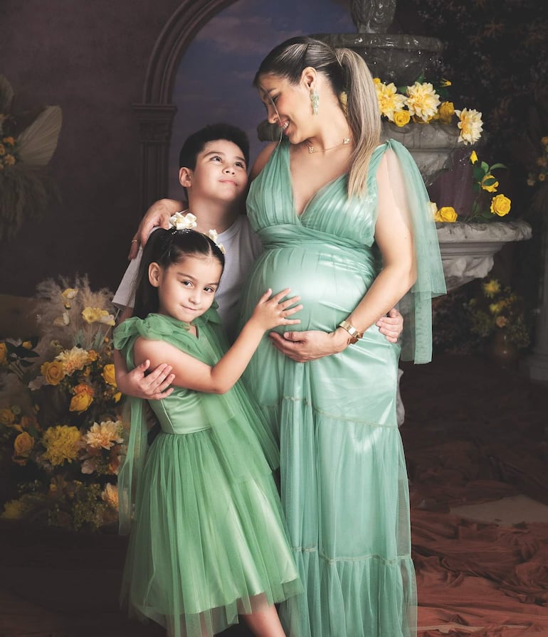 Mujer embarazada en vestido verde sonríe mientras niños la abrazan, en decoración festiva de flores amarillas.