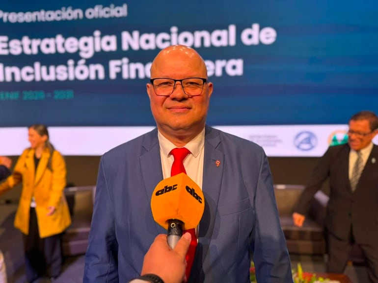 Óscar Lovera, calvo y con gafas, vestido con traje azul y corbata roja, sostiene un micrófono mientras sonríe en un auditorio.
