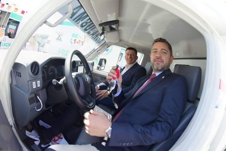 Pedro Alliana sonríe, sosteniendo llaves de ambulancia; el intendente lo observa, ambos en trajes formales dentro del vehículo.