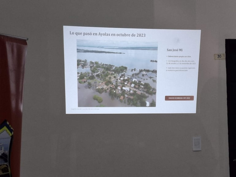 Proyección de un informe sobre inundaciones en pared, mostrando paisaje aéreo de casas inundadas en Ayolas.