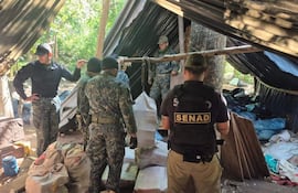 Cinco hombres con uniformes tácticos, uno con chaleco de SENAD, revisan sacos en un campamento improvisado entre árboles.