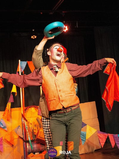 Payaso con chaleco naranja y camisa marrón sostiene pañuelo rojo, mientras otro lanza objeto azul en un ambiente festivo.