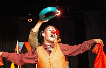 Payaso con chaleco naranja y camisa marrón sostiene pañuelo rojo, mientras otro lanza objeto azul en un ambiente festivo.