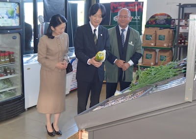 Mujer en vestido claro observa, mientras un hombre con traje oscuro sostiene un pimiento y otro con abrigo verde observa en un mercado de productos orgánicos.