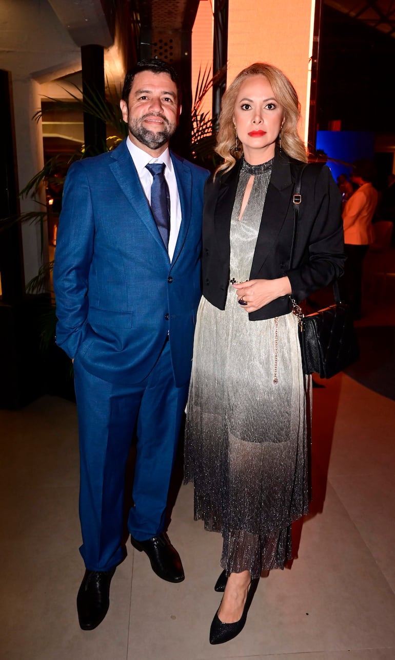 Hombre con traje azul oscuro y corbata sonríe junto a mujer en vestido plateado con abrigo negro, ambos en elegante salón.