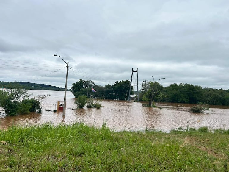 Paisaje inundado con puentes colgantes al fondo y vegetación cubierta por agua marrón en un clima nublado.