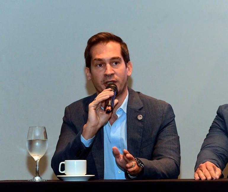 Elías Saba, en traje oscuro y camisa clara, sostiene un micrófono mientras habla, con vaso y taza en la mesa.