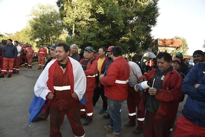 Un hombre de cabello rizado con overol rojo sostiene una bandera, rodeado de otros funcionarios en un ambiente exterior de protesta.