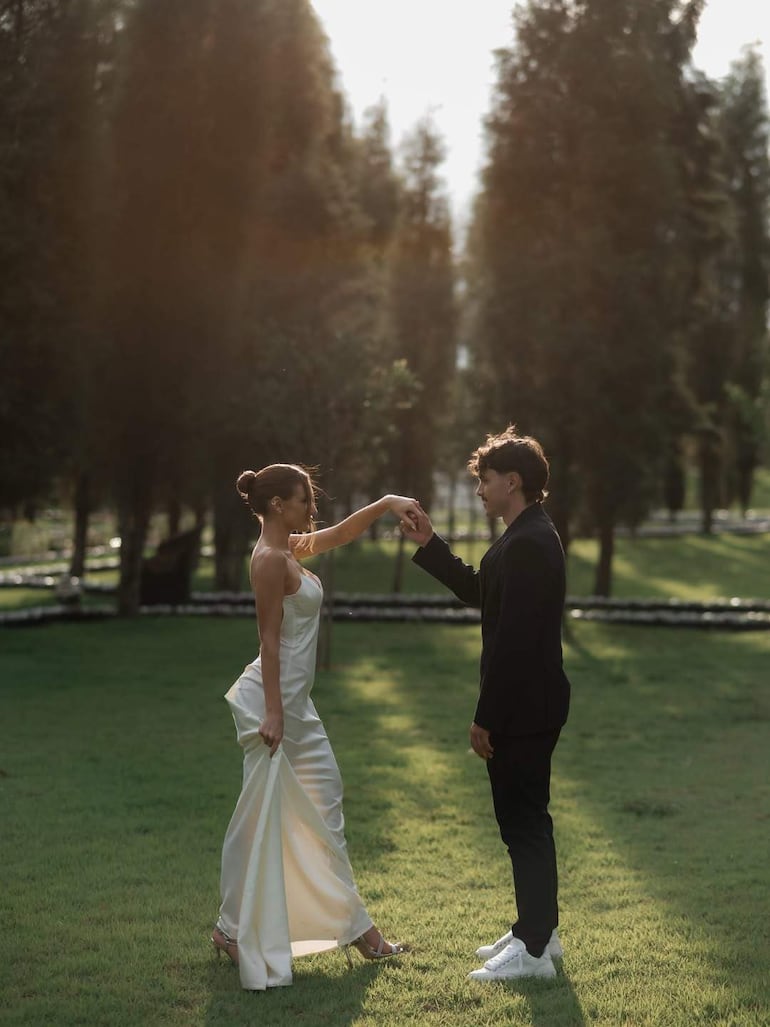 Mujer en vestido blanco y hombre en conjunto negro sostienen las manos en un entorno natural con árboles altos.