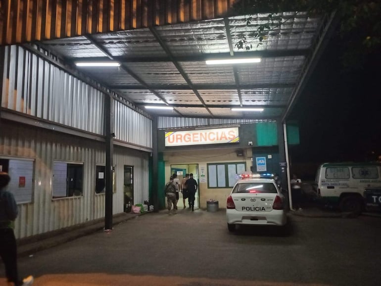 Ambiente exterior del hospital con un letrero 'URGENCIAS', vehículo policial y ambulancia visible, personas conversando.