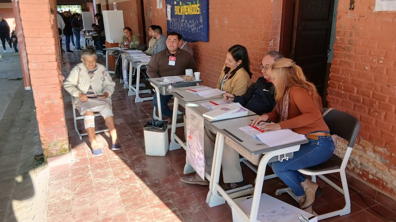 Mujer mayor con abrigo claro sentada, junto a cinco personas en mesas, en un ambiente simple de votación.