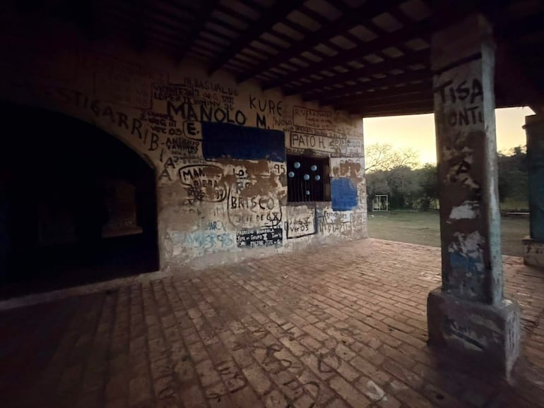 Espacio interior con grafitis y nombres escritos. Campo de fútbol visible al exterior, ambiente rural y desolado.