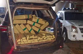 Maletero de camioneta lleno de paquetes plásticos amarillos y vehículo policial identificado como antinarcóticos en un estacionamiento oscuro.