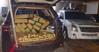 Maletero de camioneta lleno de paquetes plásticos amarillos y vehículo policial identificado como antinarcóticos en un estacionamiento oscuro.
