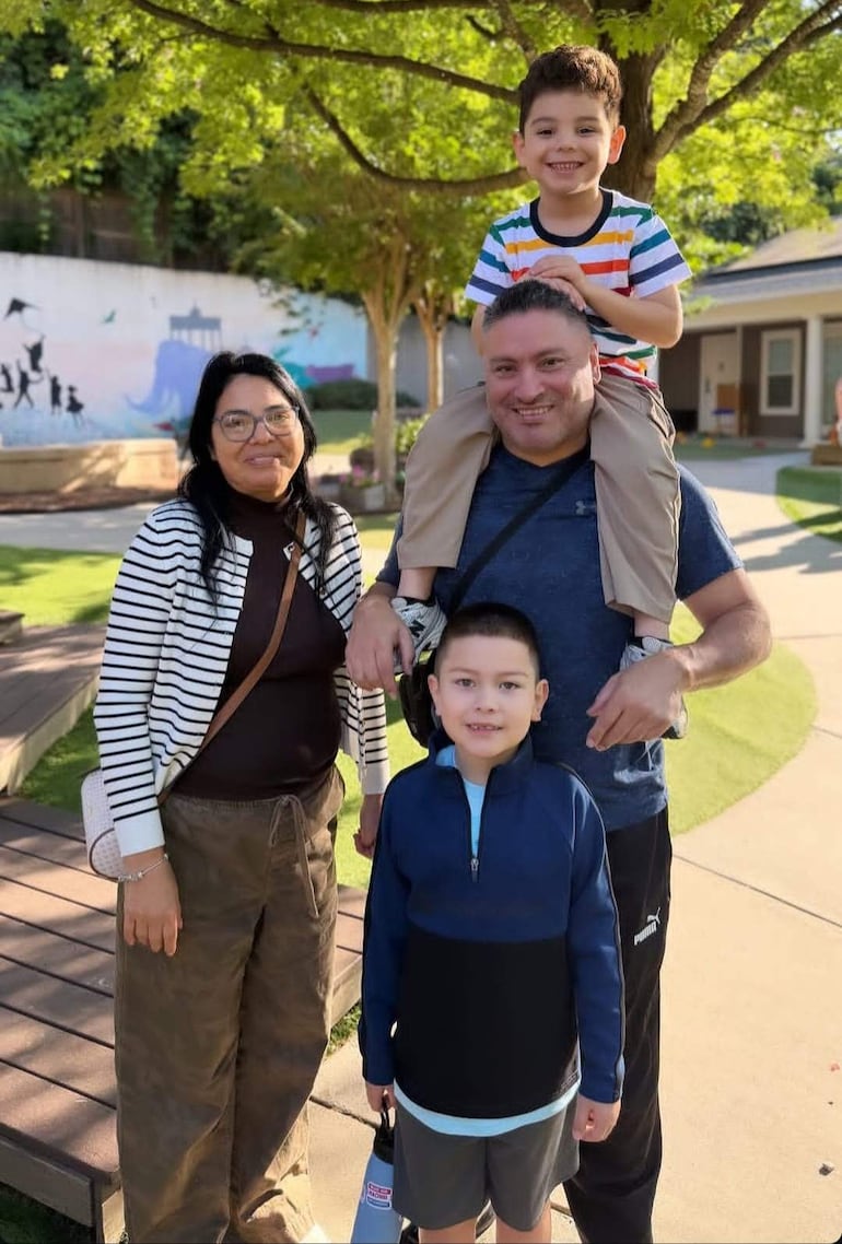 Hombre con camiseta azul sostiene a un niño sobre sus hombros, mientras una mujer y otro niño observan sonrientes en un parque.