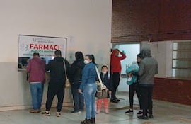 Una mujer con mascarilla sostiene una bolsa mientras observa a dos hombres conversando en el mostrador de la farmacia.