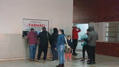 Una mujer con mascarilla sostiene una bolsa mientras observa a dos hombres conversando en el mostrador de la farmacia.