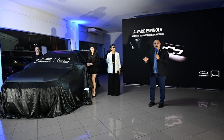 Álvaro Espínola en blazer oscuro, hablando frente a un automóvil cubierto con tela negra, dos mujeres lo observan atentamente.