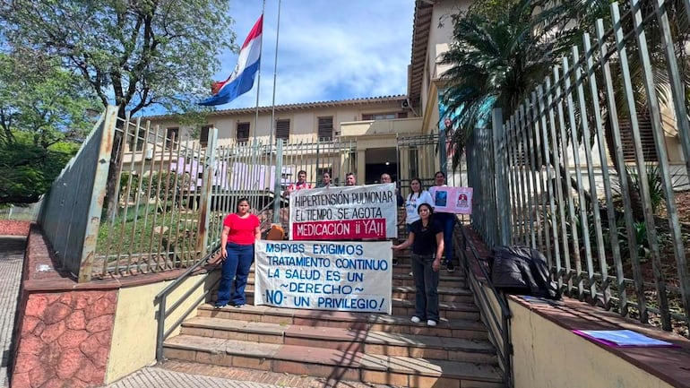 Seis manifestantes sosteniendo pancartas en protesta, expresiones tristes, ropa de jeans, con bandera de Paraguay de fondo.