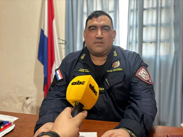 Comisario Jorge Arbo en uniforme de la Policía Nacional, sentado frente a un escritorio, sosteniendo un micrófono amarillo con expresión seria.