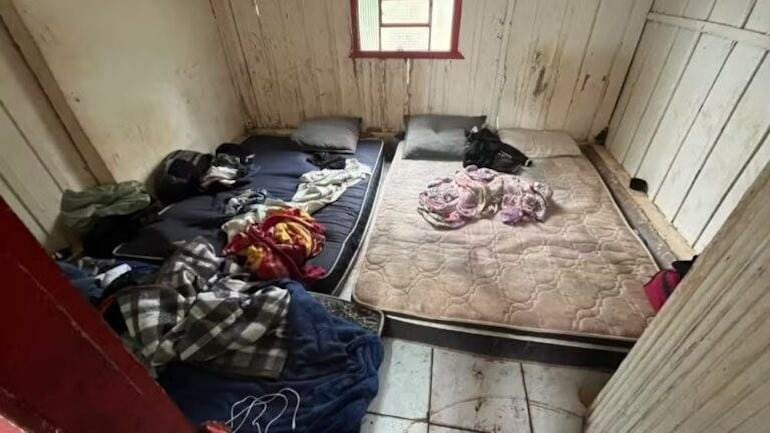 Interior desordenado con colchones en el suelo y ropa esparcida, sin personas visibles en la escena.