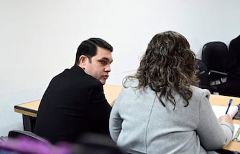 Rivas, vestido con suéter negro, escucha atentamente mientras su defensora, con abrigo gris y cabello rizado, escribe en un cuaderno.